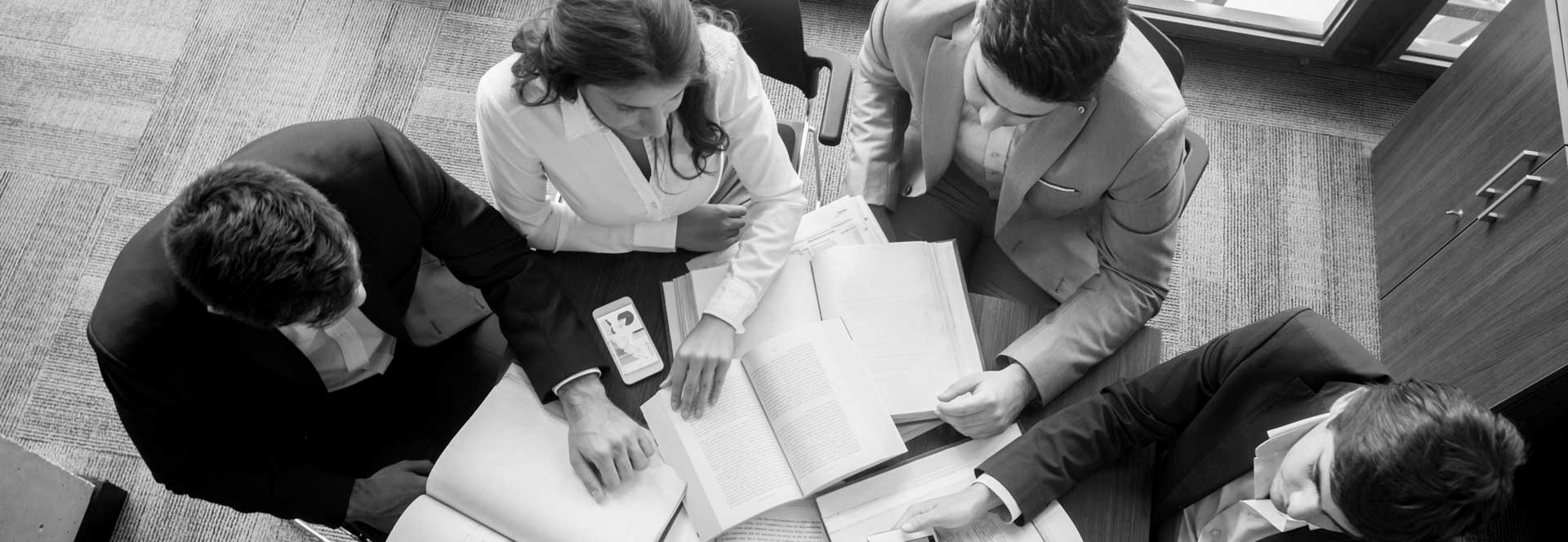 5 business people are seated at a circular table analyzing charts and data on their smartphones and tablets with 5 textbooks open